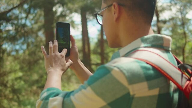 Asian man tourist taking photo on smartphone. Young hiker walking in forest camp, using mobile phone, traveling and hiking alone. Summer tourism, wild nature. 