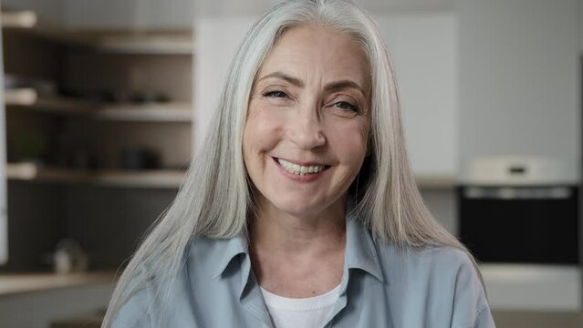 Satisfied positive gray-haired stylish old housewife mature carefree serene grandmother with long blond hair. Female portrait happy caucasian lady retiree granny wearing casual shirt posing in kitchen