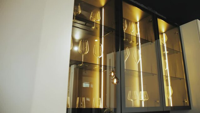 Black cupboard with glass wall in the kitchen with crockery and clear glasses with lighting. Clean wine glasses in cabinet. Contemporary Design Style. Kitchen Room. Slow motion.