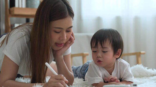 Asian young mother is teaching her cute little son to paint and drawing in the living room at home.
