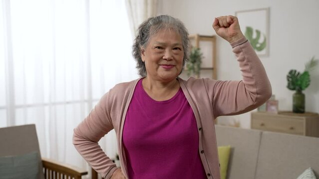 portrait of a confident asian grandmother lifting her arm in posture of power and smiling at the camera in the living room at home