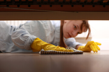 Cleaning and disinfection of surfaces. Woman with yellow rubber gloves and rag cleaning floor under bed. Beautiful woman cleaning house.