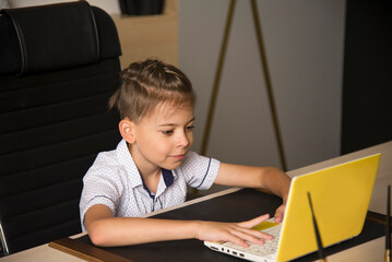 diligent schoolboy sitting at the desk with yellow laptop and studying online at school