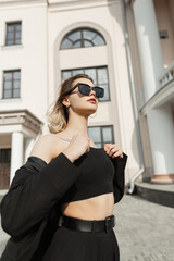 Fashionable young woman model with trendy sunglasses in black business elegant clothes walks on the street near a vintage building