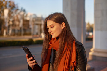 young woman using smart phone, checking news, playing mobile games or texting messages standing outdoors. Millennial lady browsing phone in city