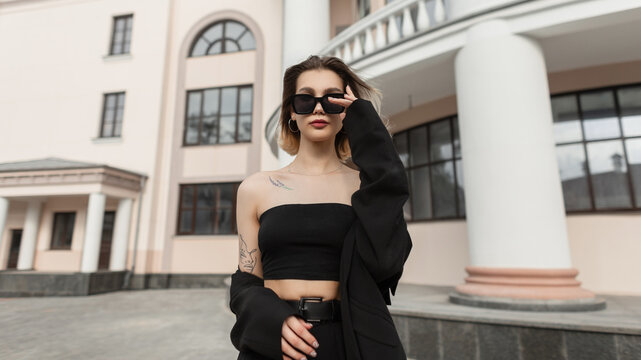 Fashionable elegant trendy woman wears trendy sunglasses in black fashion outfit with top and blazer walks in the city near a vintage building