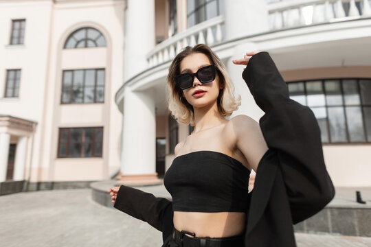 Fashionable young business woman model with stylish modern sunglasses in black fashion elegant clothes with top and blazer walks on the street near a vintage building. Urban style and fashion