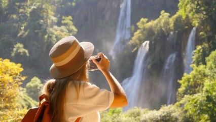 Fototapeta premium Cheerful happy joyful young woman tourist with camera makes photos for social media network or for memories on beautiful tropical waterfall. Concept of active travel, trip in Thailand