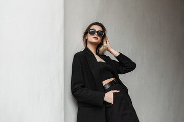 Elegant fashionable business glamour woman model with fashion sunglasses in black clothes with top and stylish black blazer stands near a wall on the street - obrazy, fototapety, plakaty