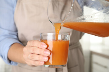 Woman pouring freshly made carrot juice into glass in kitchen, closeup