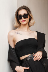 Stylish beautiful vogue elegant business model girl with fashionable sunglasses in fashion black business clothes with top and blazer standing near the white wall. Women's street portrait of woman