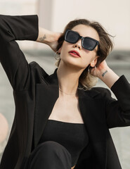 Fashionable young beautiful urban woman model with sunglasses in black fashion blazer and top sits...