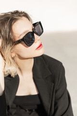Naklejka premium Summer female fresh portrait of beautiful young woman model with cool trendy sunglasses in black elegant clothes sits outdoors at sunlight