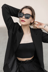 Beautiful young woman model hipster with cool modern sunglasses in elegant vogue black clothes look with blazer, top and pants on a white background on the street