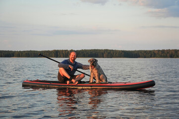 Fototapeta na wymiar A mature man with a dog is paddling on the lake water on a SUP board. Vacation, tourism, active lifestyle.