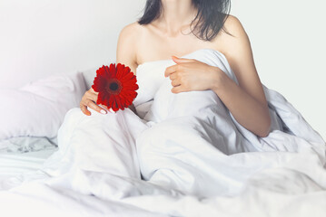 Obraz na płótnie Canvas young woman on a gentle morning in bed with white bed linen with a red flower in her hands. close-up