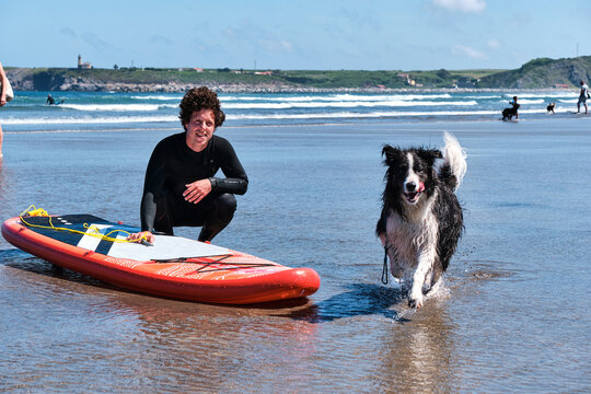 Young surfer with long curly hair posing with his paddle surf board and his border collie dog while it is running