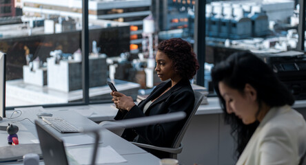 African American female employee using her smartphone at her desk in the office located in a skyscraper. Modern friendly working environment