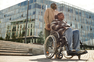 Low angle portrait of smiling black man assisting partner in wheelchair and enjoying walk together in city against glass architecture, copy space