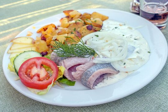 Pickled herring or matjes fillet with cream sauce and onions, served with fried potatoes and salad on a white plate, copy space, selected focus