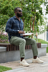 Vertical full length portrait of blind man wearing sunglasses and reading book in braille outdoors