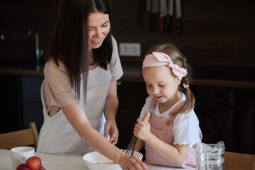 Mother and daughter preparing a sweet cake using flour, milk, sitting on chairs at a table in a...