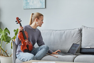 Side view portrait of young woman playing violin at home and composing music with laptop, copy space