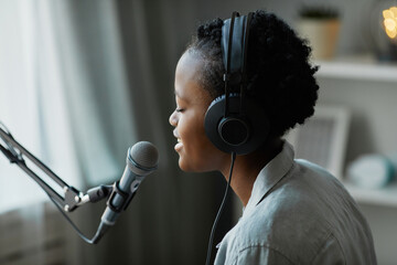 Minimal side view of young female musician singing to microphone at home and wearing headphones,...