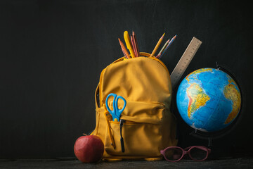 Yellow backpack with school supplies next to the globe, red apple and glasses on the black school...