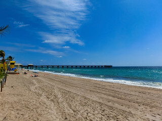 Long distance view of the sunny and tropical blue water shoreline with people, foliage, boats and buildings in the background seen from above the water.