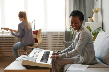 Side view portrait of smiling black woman playing synthesizer at home and composing music in...