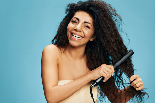 Enjoyed Latin curly woman using hair straightener, looking aside, posing isolated on blue wall background. Hair routine concept, haircare, dry damaged hair ironing, hairdressing offer, copy space