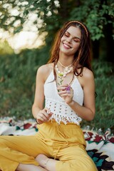 Young hippie woman sitting in nature on a plaid with a flower in her hands and looking at the camera smiling sincerely in eco-clothing in autumn in the sunset