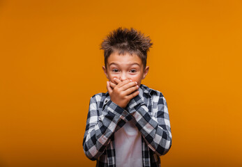 childhood, expressions and the concept of people - a shocked little boy covering his mouth with his hands on a yellow background