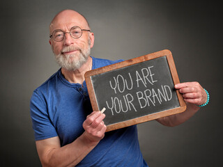 senior man, businessman or mentor, is showing a blackboard  with you are your brand sign in white chalk, branding and marketing concept