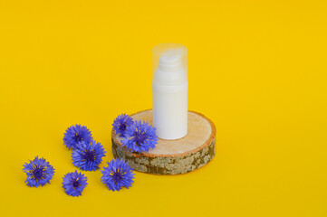 A white plastic jar for cosmetics stands on a wooden podium with blue cornflowers on an yellow background.
