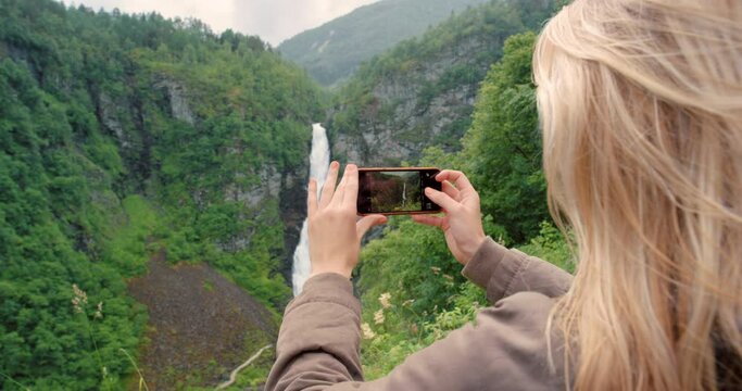 A nature tourist taking photos on phone of the green mountain, waterfall view on hiking adventure outdoors. Young activist or environmentalist showing eco friendly environment on social media
