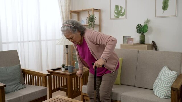 unwell asian grandmother moving around with cane walking stick at home is sitting down to rest on the couch and rubbing her painful back in agony at daytime in the living room