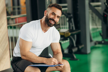 Muscular handsome trainer looking at fitness plan on clipboard for working out in the fitness gym