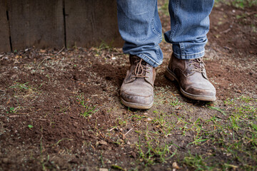 Man's feet and lower legs in blue denim jeans and brown work man's boots standing on patchy garden lawn. 