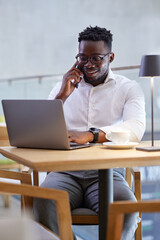 An African businessman multitasks while sitting in a cafe. A businessman types on the laptop and talks on the phone with a client.