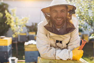 Portrait of a happy male beekeeper working in an apiary near beehives with bees. Collect honey. Beekeeper on apiary. Beekeeping concept. - obrazy, fototapety, plakaty