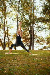 A fit woman is doing yoga outside. She is in the warrior yoga posture.