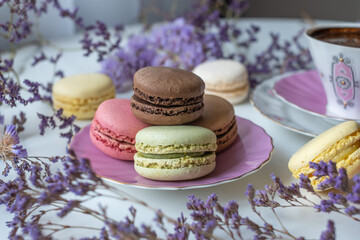Obraz na płótnie Canvas Mix of appetizing macarons and delicious coffee in porcelain cup on white table decorated with blurred flowers. Dry flowers. Greeting card.