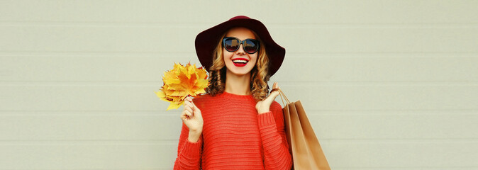 Autumn portrait of happy smiling young woman with shopping bags and yellow maple leaves wearing sweater and hat on gray background