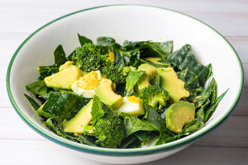 Avocado salad with broccoli, boiled egg, spinach in white vintage bowl on light salad