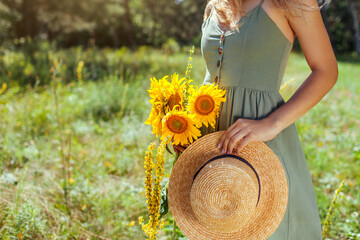 Close up of summer female accessory and clothes. Woman holding straw hat with bouquet of sunflowers...