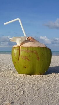 Vertical footage of coconut juice with a white straw on the beach in sunlight