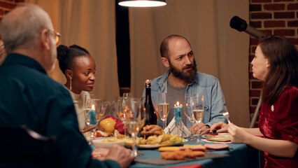 Joyful multiethnic people in dining room celebrating winter holiday with traditional home cooked food. Happy festive family enjoying Christmas dinner at home while eating together. Handheld shot