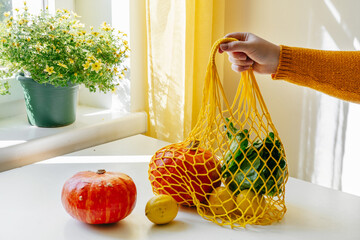 Female hand holding shopping net with vegetables - pumpkins and green salad - at the kitchen. Yellow and orange colors. Zero waste concept